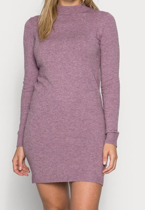 Femme portant une robe en maille mauve ajustée à manches longues avec un col montant, debout devant un fond clair uni.