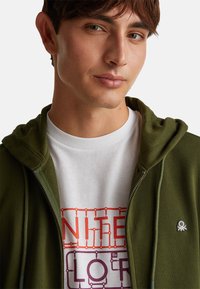 Felpa con zip di colore verde oliva con una texture morbida, abbinata a una t-shirt bianca con testo grafico colorato. Piccolo logo sulla felpa.