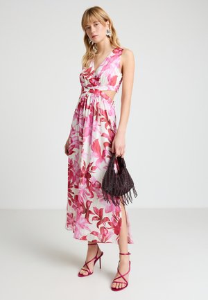 Femme portant une robe longue fleurie rose et rouge avec des découpes, tenant un sac à main noir orné de perles et portant des talons roses à brides assortis.