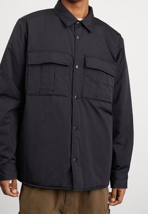 Mann trägt eine schwarze Jacke mit Knöpfen, zwei großen Klappentaschen auf der Brust und braune Hosen, steht vor einem einfarbigen Hintergrund.