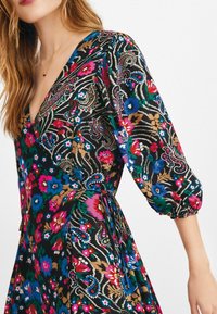 Robe fleurie colorée avec un fond noir, des motifs vibrants en rose, bleu et vert, un décolleté plongeant en V, et des manches bouffantes avec un détail noué.