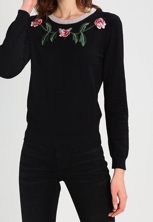 Zwarte trui met geborduurde rode rozen en groene bladeren langs de halslijn, gedragen met zwarte broek door een staand persoon.