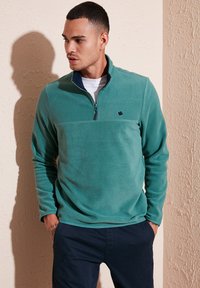 Pullover in pile color turchese con mezza zip e colletto blu navy. Presenta un piccolo logo sul petto e maniche lunghe con vestibilità comoda.