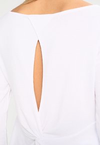Top bianco a maniche lunghe con scollatura ampia, taglio a chiave sulla schiena e dettaglio attorcigliato sull'orlo per un look aderente. Tessuto testurizzato.