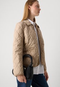 Kvinde i beige quiltet jakke og hvid skjorte bærer en sort lædertaske og står sidelæns mod en ensfarvet baggrund.