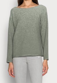 Vihreä pitkähihaisessa paidassa on teksturoitu pinta, leveä kaula-aukko ja hieman lyhyt helma. Yhdistetty vaaleanharmaisiin farkkuihin.