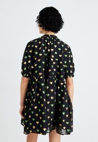 Svart kjole med pufferm, prydet med grønne og gyldne hjerteformer, strukturert stoff og rynket halslinning, designet i en løs silhuett.