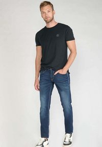 T-shirt en coton noir, jean slim bleu et sneakers noir et blanc. Un mannequin masculin se tient face à l'avant sur un fond clair.