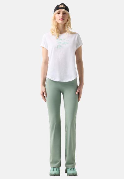 Junge Frau, die ein weißes T-Shirt mit Grafik, salbeigrüne Hose, mintgrüne Schuhe und eine schwarze Baseballkappe rückwärts trägt, vor einem einfarbigen Hintergrund steht.