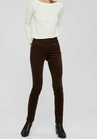 Femme portant un haut à manches longues blanc, un pantalon slim marron foncé et des bottes noires à la cheville sur un fond gris uni.