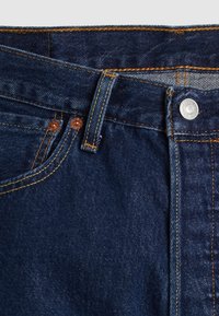 Primer plano de unos jeans de mezclilla azul oscuro que muestra la cinturilla, el lazo del cinturón, el botón, los remaches y los detalles de costura en color naranja.