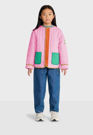 Enfant portant une veste matelassée rose avec des poches vertes, un jean bleu et des baskets blanches, debout devant un fond clair uni.