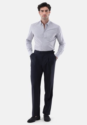 Mann steht mit Händen in den Taschen, trägt ein kariertes Hemd mit langen Ärmeln, dunkle maßgeschneiderte Hosen und schwarze Loafers auf weißem Hintergrund.