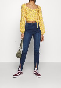 Gul blommig croptopp med puffärmar, knuten i midjan; mörkblå högmidjade jeans med sidospjäll; vinröda sneakers; grön handväska.