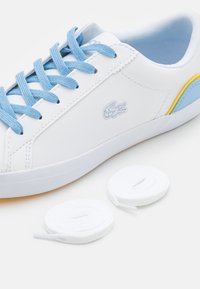 White sneakers with blue laces and accents. Features a smooth synthetic upper, rounded toe, and a small embroidered logo. Includes spare white laces.