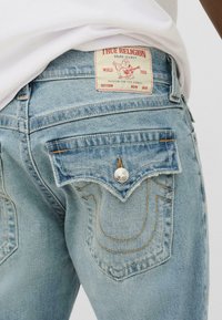 Svijetloplave traperice s izblijedjelim završetkom, s oznakom True Religion i karakterističnim zakrivljenim šavovima na stražnjim džepovima.