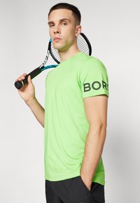 Zöld sportpóló rövid ujjakkal, a bal ujján "BORO" felirattal, amelyet egy teniszütőt tartó személy visel.