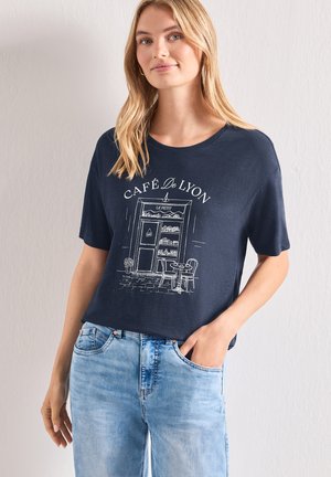 Lyshåret kvinde iført en mørkeblå t-shirt med en hvid illustration af Café de Lyon's facade og lyseblå jeans med den ene hånd i lommen.