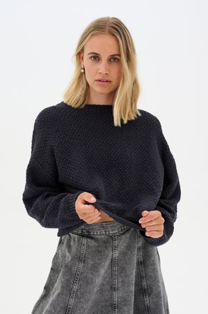 Marineblauwe gebreide trui met een gestructureerd patroon, hoge halslijn en verlaagde schouders, gecombineerd met een grijze denim rok met paneelnaden.