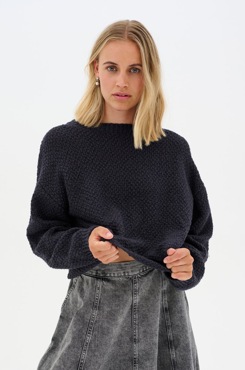 Pull en maille bleu marine avec un motif texturé, un col montant et des épaules tombantes, associé à une jupe en denim gris présentant des coutures de panneaux.