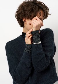 Persoon met kort krullend haar, gekleed in een donzige donkere trui, met een zilveren armband, ring en oorcuffs, houdt hun gekruiste handen dicht bij hun gezicht.