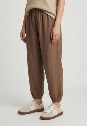 Osoba nosząca luźne brązowe spodnie z elastycznymi mankietami i beżowe trampki z białymi sznurowadłami, stojąca na jednolitym jasnym tle.