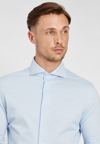 Camicia azzurro chiaro con bottoni, realizzata in tessuto liscio, caratterizzata da un colletto classico e una leggera lucentezza, con bottoni bianchi lungo la parte frontale.