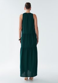 Žalia ilgų suknelių be rankovių su sluoksniuota konstrukcija, turinti susmulkintą kaklo liniją ir subtilią tekstūrą. Baigta užsegimu su skylute nugaroje.