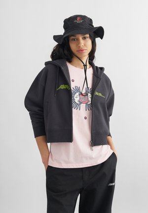 Νεαρός με μαύρο καπέλο και φούτερ Santa Cruz, ροζ γραφικό μπλουζάκι και μαύρο παντελόνι, με τα χέρια στις τσέπες, στέκεται μπροστά σε μονόχρωμο φόντο.