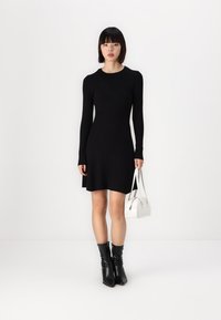 Robe en tricot côtelé noire avec des manches longues, corsage ajusté et ourlet évasé ; associée à un petit sac à main blanc et des bottines noires.