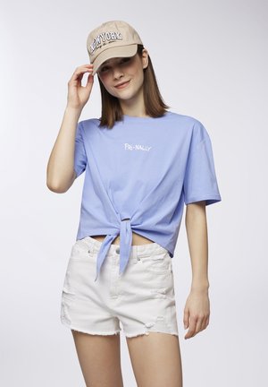 T-shirt corta azzurro chiaro con nodo frontale, con la scritta bianca "FRI-NALLY." Abbinato a pantaloni corti bianchi strappati e un cappellino beige.