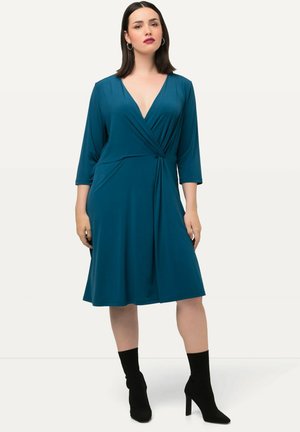 Teal, polvipituinen kietaisumekko, jossa on 3/4-pituiset hihat, laskostettu kangas ja kiemurteleva yksityiskohta vyötäröllä, yhdistettynä mustiin nilkkasaappaisiin.