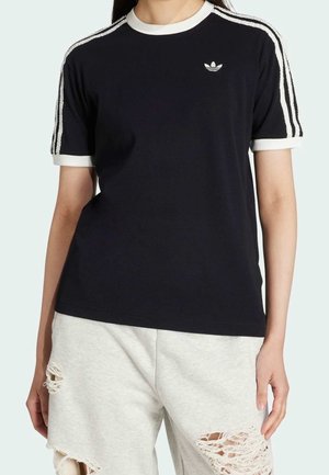 Persoon draagt een zwart Adidas T-shirt met witte strepen op de mouwen en een klein logo, gecombineerd met lichtgrijze gescheurde korte broek.