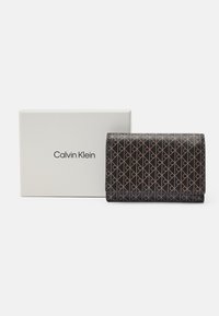 Peňaženka Calvin Klein v čiernom textúrovanom materiáli s opakujúcim sa vzorom loga a béžovými akcentmi, vystavená vedľa bielej krabičky s logom.