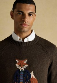 Ein junger Mann trägt einen braunen Pullover mit einem detaillierten Fuchsdesign in einer blauen Jacke über einem weißen Hemd vor einem beigen Hintergrund.