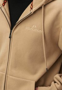 Beżowa bluza z kapturem na zamek błyskawiczny z polarem wewnątrz, z wyszywanym logo, pomarańczowymi i czarnymi sznurkami w wzór oraz kieszeniami z przodu.