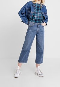 Denim jacket in dark blue, retracted sleeves. Green plaid short-sleeve top. Light blue wide-leg jeans. White high-top sneakers.
