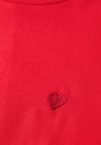 Punainen kangas, jossa on pieni, kirjailtu sydämen muoto hieman tummemman punaisella langalla ommeltuna lähellä keskustaa.