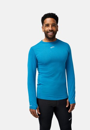 Langärmliges Sportshirt aus hellblauem Stoff, mit rundem Ausschnitt, figurbetontem Schnitt und Logo auf der Brust, kombiniert mit schwarzen Hosen.