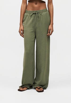 Άτομο που φοράει χακί παντελόνι φαρδιάς γραμμής με ελαστική μέση και κορδόνι, σε συνδυασμό με μαύρα σαγιονάρες, στέκεται μπροστά σε λευκό φόντο.