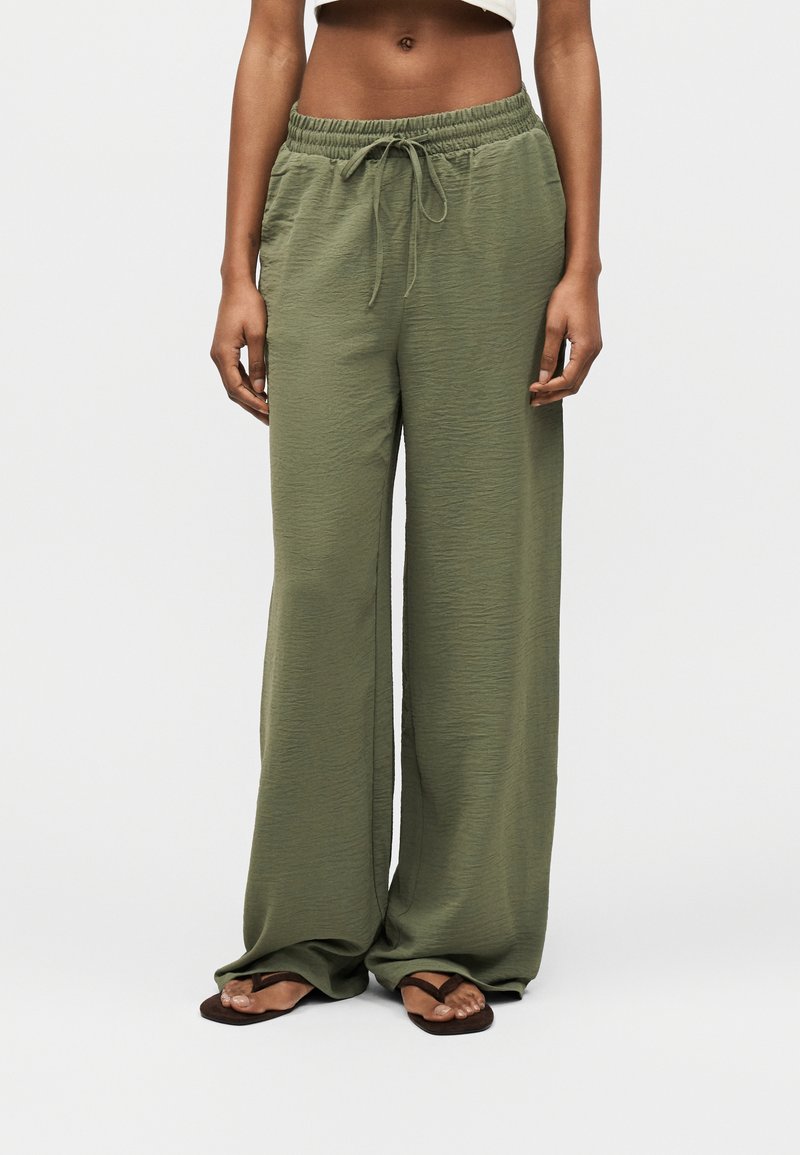 Άτομο που φοράει χακί παντελόνι φαρδιάς γραμμής με ελαστική μέση και κορδόνι, σε συνδυασμό με μαύρα σαγιονάρες, στέκεται μπροστά σε λευκό φόντο.