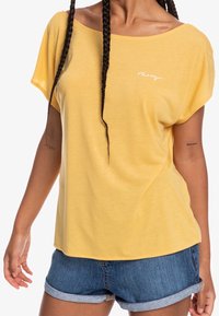 Keltainen lyhythihainen paita, jossa on väljä malli ja leveä kaula-aukko, jossa on pieni valkoinen logo. Yhdistettyinä farkkushortseihin, luoden rentoa tyyliä.