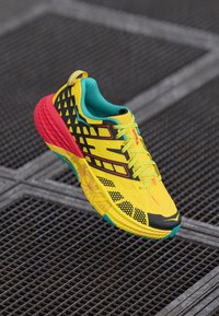 Baskets de course jaune vif avec un design texturé noir et rouge, des accents turquoises et des détails réfléchissants sur les lacets et la semelle.