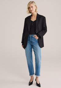 Čierne sako cez čierne tričko, v kombinácii so svetlomodrými džínsami s vyhrnutými manžetami a čiernymi topánkami so špicatou špičkou. Hladká látka a prispôsobený strih.