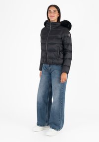 Giubbotto trapuntato nero con cappuccio foderato in pelliccia, dotato di zipper frontale, design leggermente corto e cuciture visibili, abbinato a jeans a gamba larga.