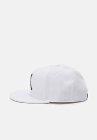 Hvid bomulds baseball cap med flad skygge og broderet sort logo. Har syningsdetaljer og ventilationshuller på kronen.