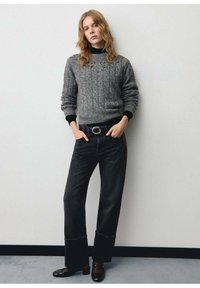 Maglione grigio a maglia con decorazioni, abbinato a jeans neri a vita alta e scarpe nere. Sfondo semplice.