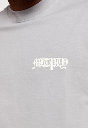 Mann trägt hellgraues T-Shirt mit weißem gotischem Schriftzug „MLTPLY“ auf der linken Brust.