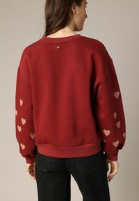 Sweatshirt rouge à coupe décontractée, manches longues avec broderie de cœur doré, col côtelé, et détail au dos avec un petit bouton.