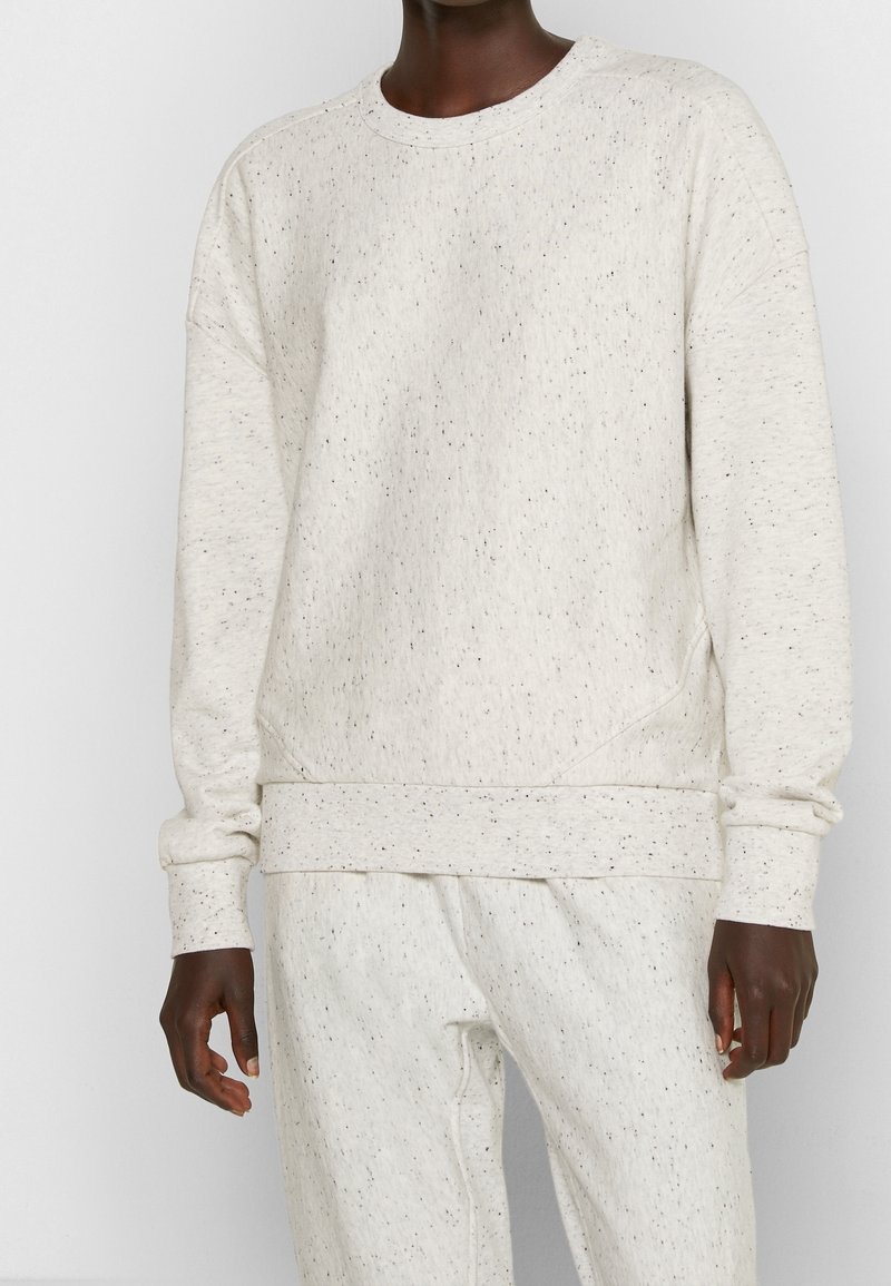 Personne portant un sweat-shirt beige clair tacheté et un pantalon assorti, debout devant un fond blanc uni.